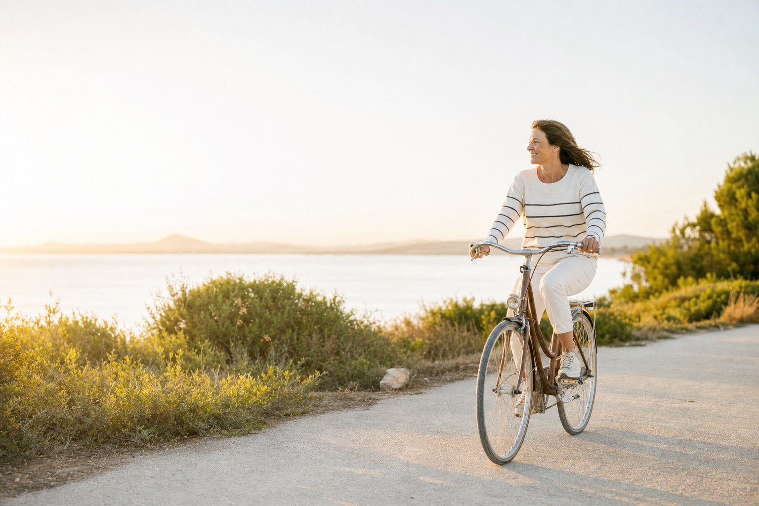 Mujer adulta montando en bicicleta junto al mar con luz cálida de atardecer.